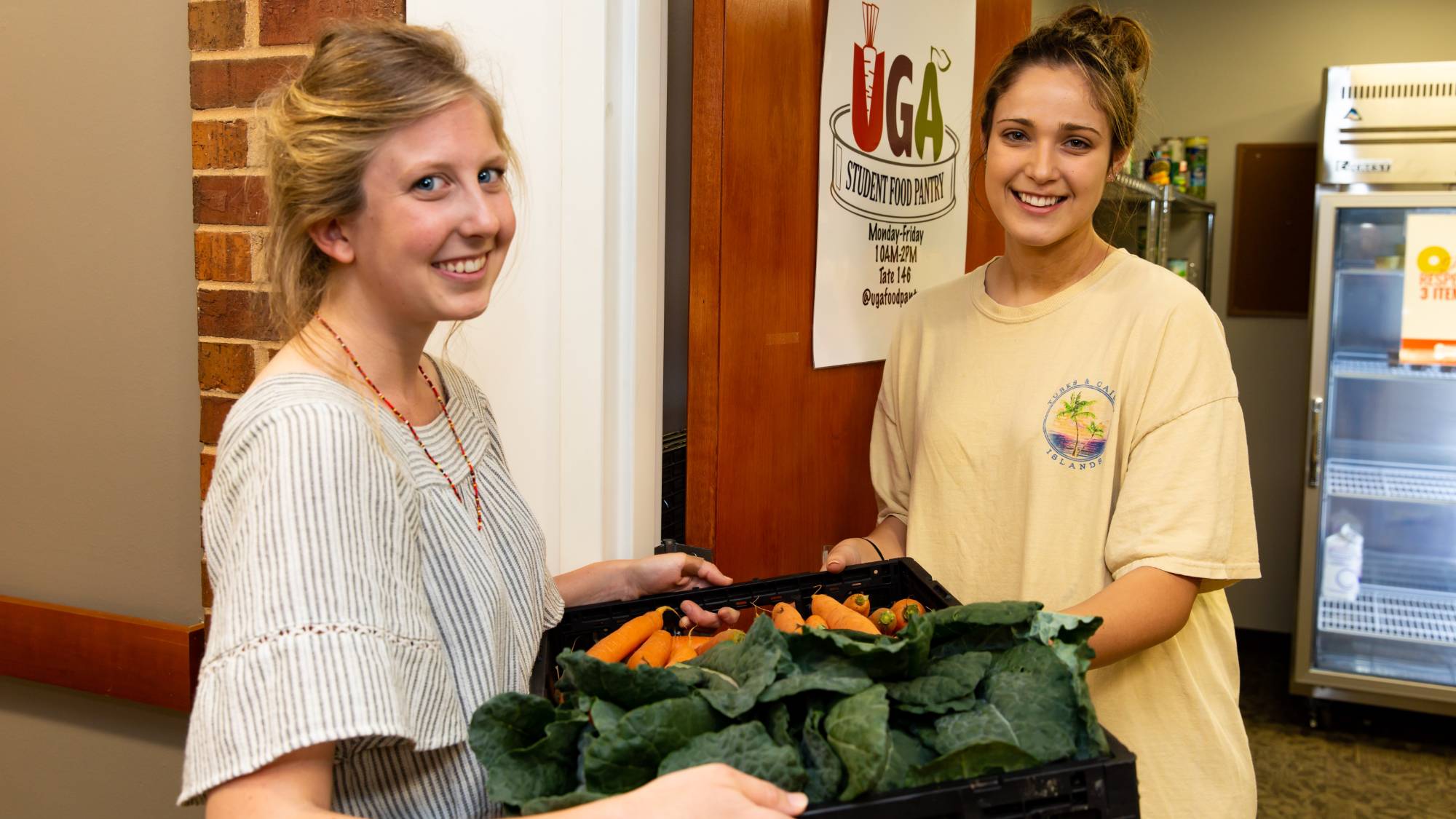 Two young women smile while one hands a tray of fresh vegetables to the other in a doorway labeled UGA Food Pantry
